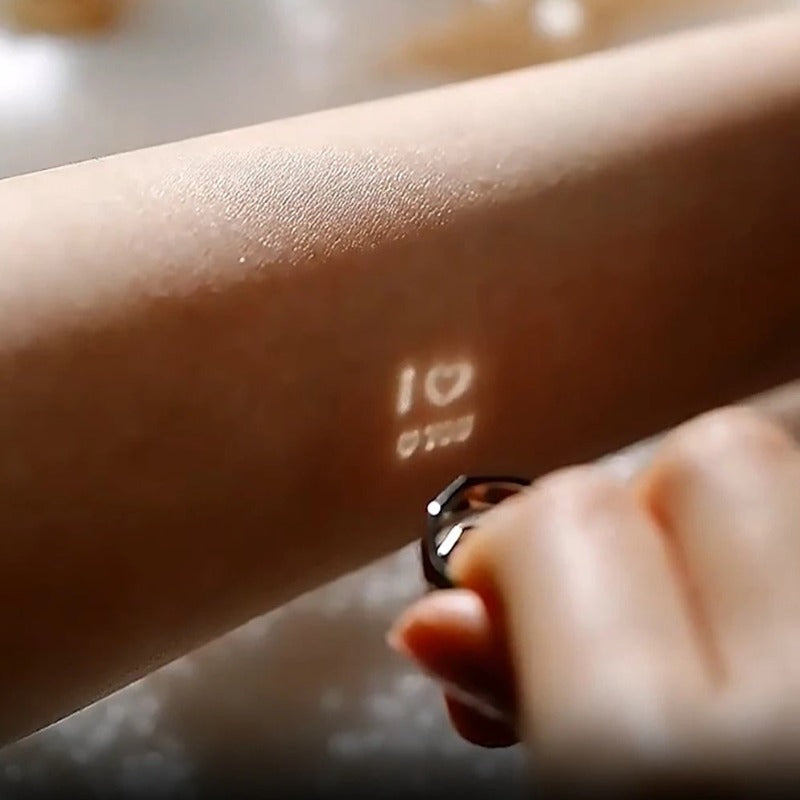 A hand holds an adjustable ring close to a forearm. The Jingqian S925 Sterling Silver Light Reflection Ring projects a small light that displays "I ❤️ U" and another small symbol or text below it on the skin.
