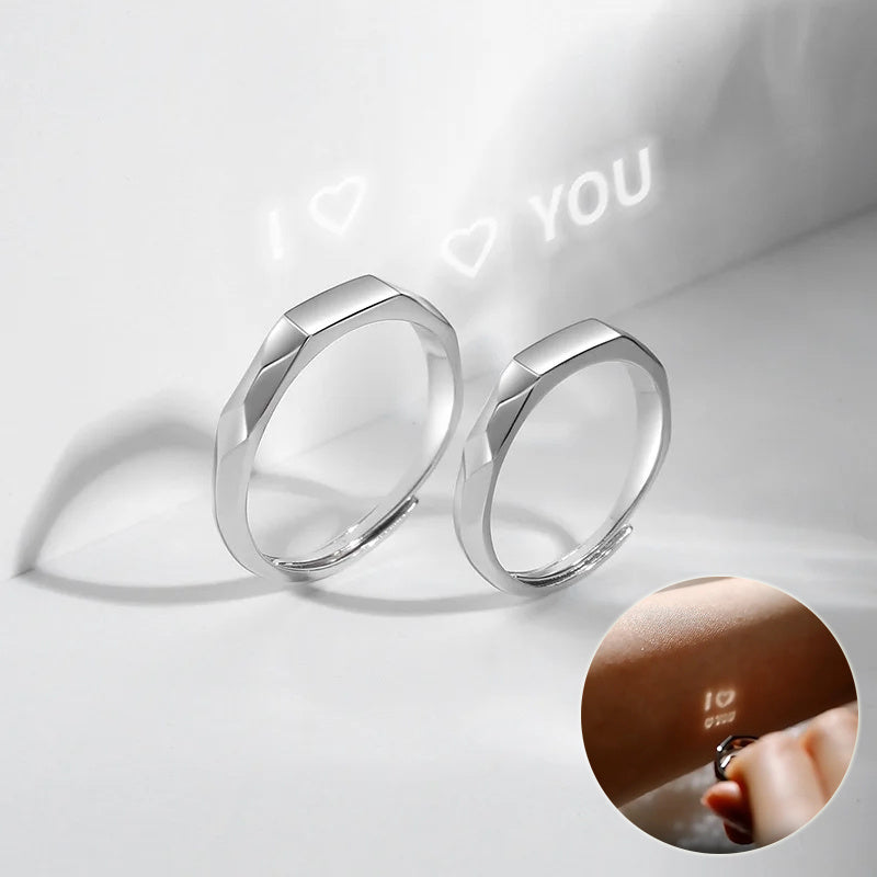 Two sterling silver rings with a flat, angular design are shown. One ring projects "I ♥ YOU" in light onto a surface. An inset image displays a close-up of someone holding an adjustable Jingqian S925 Sterling Silver Light Reflection Ring and the same "I ♥ YOU" message projected on their finger.