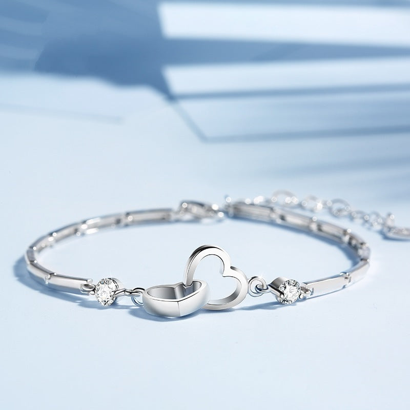 A 925 Sterling Silver Charms Bracelet with a delicate design, featuring two intertwined heart charms as the central focal point. The Jingqian charm bracelet is also adorned with small, sparkling gemstones. The background is softly lit with shadows from a nearby window.