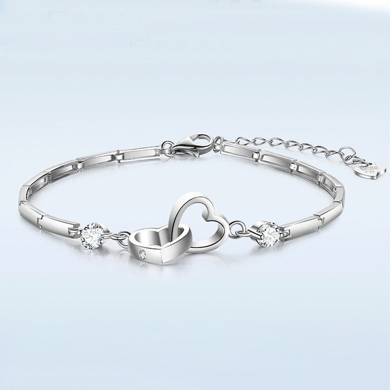 A Jingqian 925 Sterling Silver Charms Bracelet featuring two interlocking heart charms in the center, adorned with sparkling gemstones on either side. The bracelet has a sleek, linked design and an adjustable chain with a clasp closure. The background is a smooth gradient of light blue.