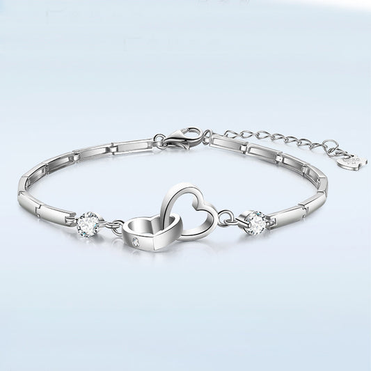 A Jingqian 925 Sterling Silver Charms Bracelet featuring two interlocking heart charms in the center, adorned with sparkling gemstones on either side. The bracelet has a sleek, linked design and an adjustable chain with a clasp closure. The background is a smooth gradient of light blue.