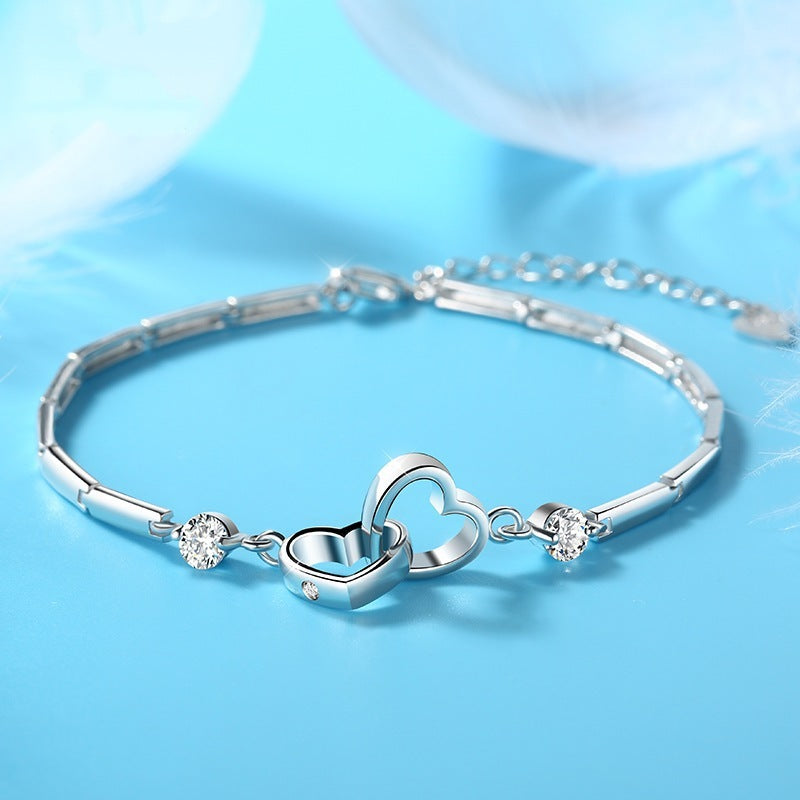 A delicate charm bracelet featuring two interlocked heart-shaped charms at the center, accented by small, sparkling crystals on both sides. The Jingqian 925 Sterling Silver Charms Bracelet is displayed on a light blue surface with white feather accents, enhancing its elegant appearance.