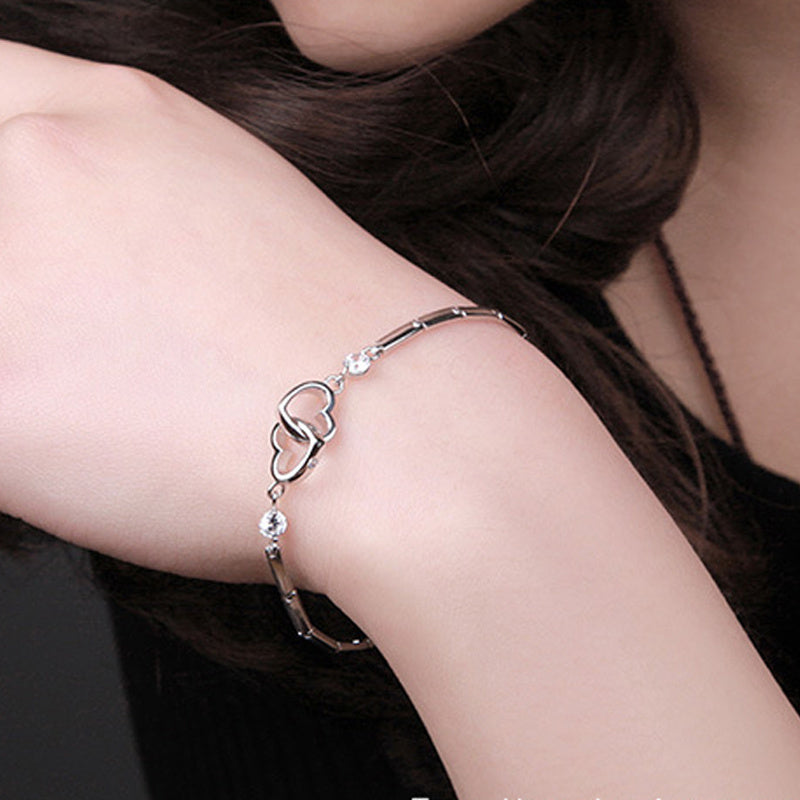 A close-up image of a person's wrist adorned with Jingqian's delicate 925 Sterling Silver Charms Bracelet. The charm bracelet features a hollow heart charm and several small round stones. The person's skin tone is light, and they have dark hair.