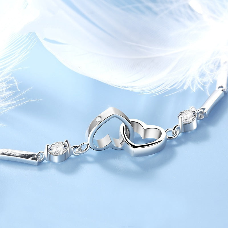 A delicate charm bracelet is shown on a light blue background with white feathers. The centerpiece consists of two interlocking heart-shaped charms, flanked by small, round crystal accents embedded in silver settings. This Jingqian 925 Sterling Silver Charms Bracelet exudes elegance and romance.