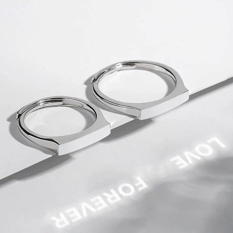 Two sleek sterling silver rings with polished surfaces are shown. One ring lays flat while the other stands angled against a white background. Light reflects off their surfaces, casting soft shadows. The words "LOVE FOREVER" are illuminated and reflected below the adjustable rings. The rings, known as S925 Sterling Silver Light Reflection Ring, are crafted by Jingqian.