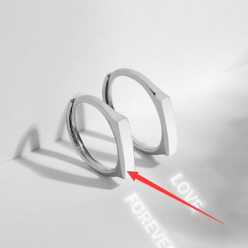A pair of S925 Sterling Silver Light Reflection Ring by Jingqian with a modern, angular design sit on a white surface. A red arrow points to the bottom ring, which casts a light reflection revealing the words "LOVE FOREVER" on the surface. The background is softly lit with gentle shadows.