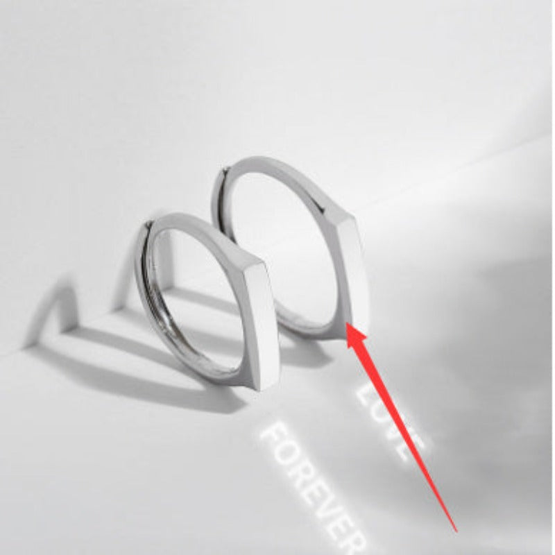 Two S925 Sterling Silver Light Reflection Rings from Jingqian with square edges are placed on a white surface. A red arrow points to one ring, with the words "LOVE" and "FOREVER" faintly visible on the surface in the background. Shadows cast by the rings and light reflections highlight their elegance.