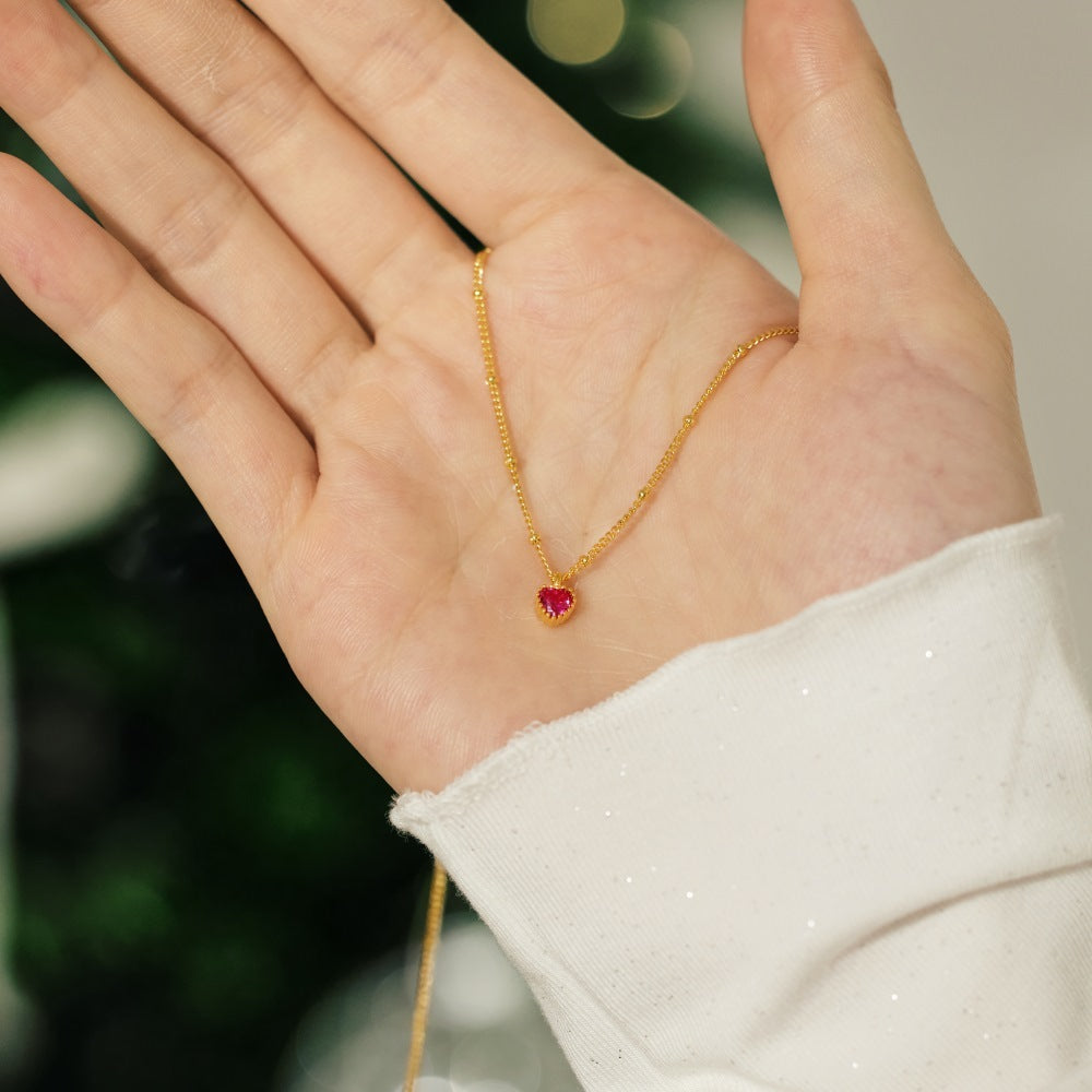 Gold necklace with a red heart pendant held in a hand against a blurred background