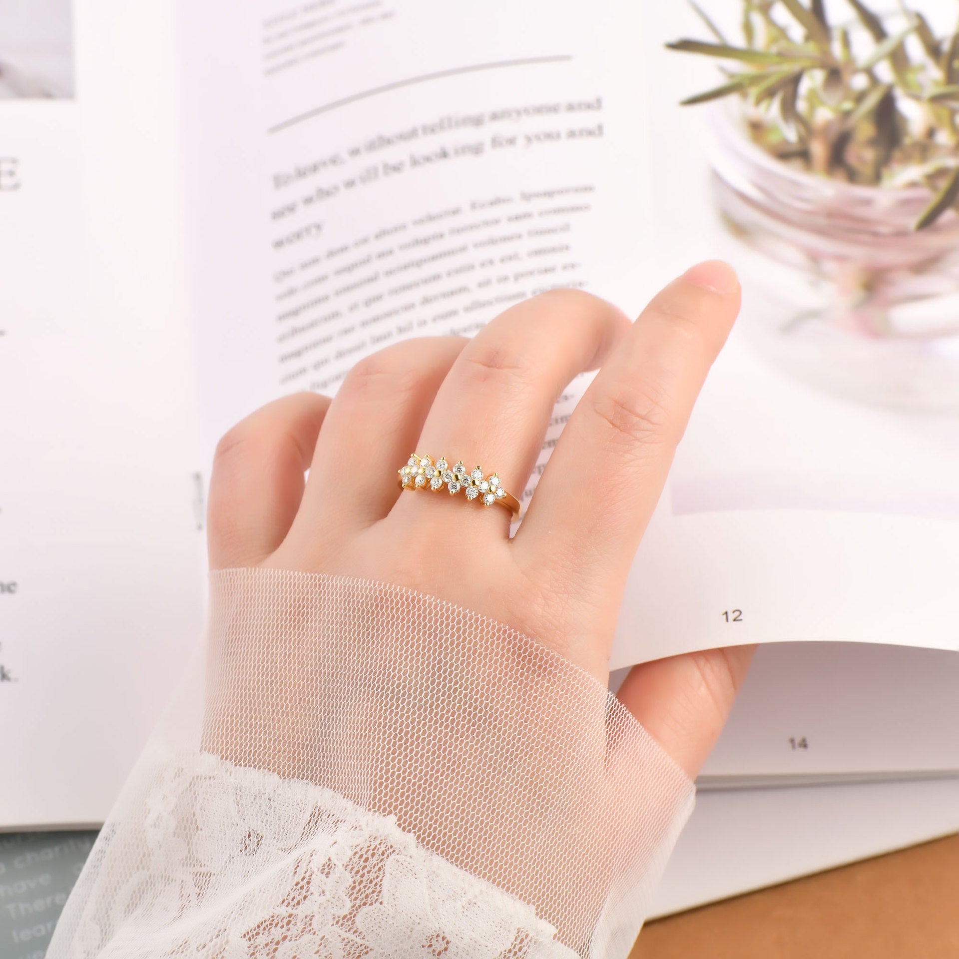 Hand wearing a gold ring with a blurred book and plant in the background